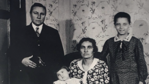 Das alte schwarz-weiß Bild zeigt eine Familie. Links eine männlich gelesene Person in Anzug. In der Mitte eine weiblich gelesene Person in einem Kleid mit Baby auf dem Arm, rechts ein etwas älteres Kind.