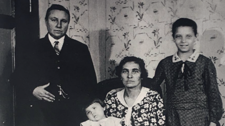 Das alte schwarz-weiß Bild zeigt eine Familie. Links eine männlich gelesene Person in Anzug. In der Mitte eine weiblich gelesene Person in einem Kleid mit Baby auf dem Arm, rechts ein etwas älteres Kind.
