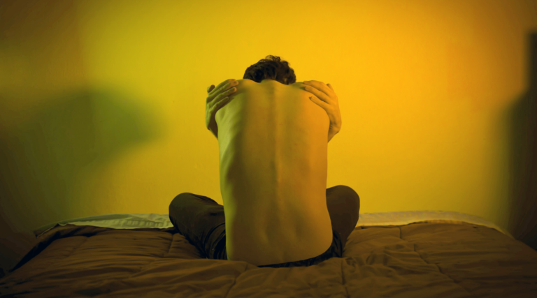 Das Bild hat eine düstere Stimmung und zeigt den nackten Rücken eines jungen Mannes der zusammengekauert auf einem Bett sitzt und sich selbst eng umarmt.